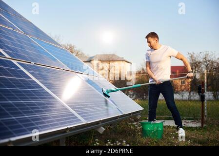 Young man is cleaning a solar panel with a mop, pv plant in rural area, side view Stock Photo