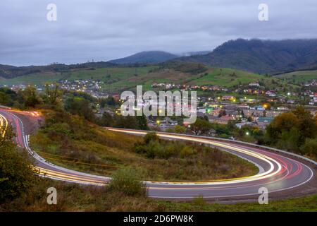 Cloudy evening on a mountain road. Headlight trails of fast moving cars. A small village shines with lights in the valley Stock Photo