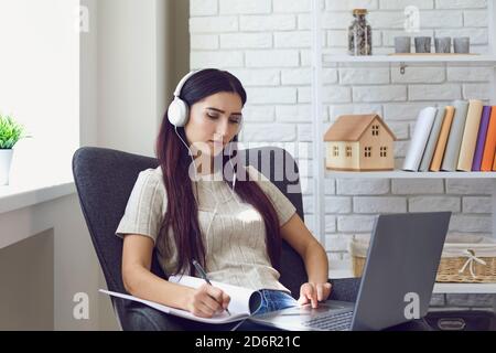 Woman in headphones and making notes during online lesson at home on laptop Stock Photo