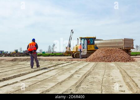 View on construction worker, supervisor, with safety vest and helmet gives instructions to workers in bulldozer and truck driver after unloading fract Stock Photo