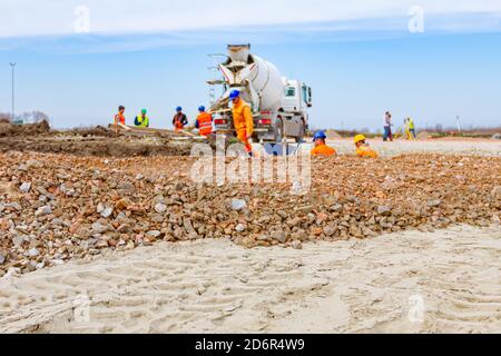 Mixer truck is unloading fresh concrete in trench, pouring over ramp. Workers are working at building site. Stock Photo