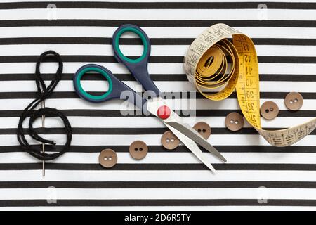 Scissors, measuring tape, needle, threads and buttons in the form of notes on striped fabric. Stock Photo