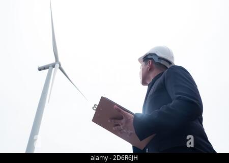 Male engineer looking and writing plan on clipboard against wind turbine Stock Photo