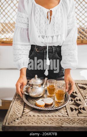 Woman hands holding silver tray with traditional moroccan mint tea, cookies and vintage teapot. Hospitality and service in Morocco. Stock Photo