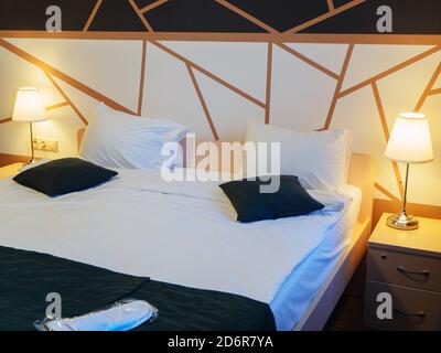 A neat double bed with lit floor lamps on the nightstands and a geometric pattern on the wall. Hotel room Stock Photo