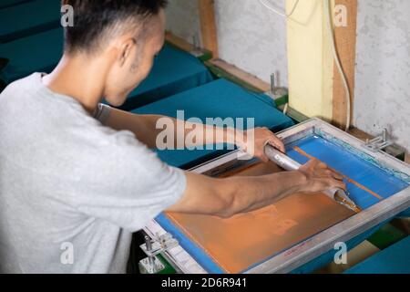Young confident, experienced male working with squeegee on a small factory, handsome worker man using printmaking tools, screen printing on clothing fabric technique Stock Photo