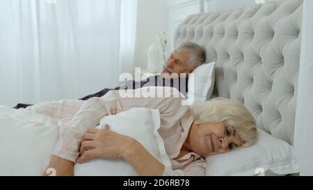 Senior Caucasian grandparents couple lying and sleeping in bed at home. Irritated woman getting disturbed with man loudly snoring Stock Photo