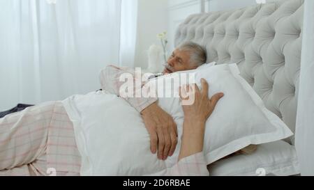 Senior Caucasian grandparents couple lying and sleeping in bed at home. Irritated woman getting disturbed with man loudly snoring. Grandmother covering her ears with pillow while grandfather snores Stock Photo