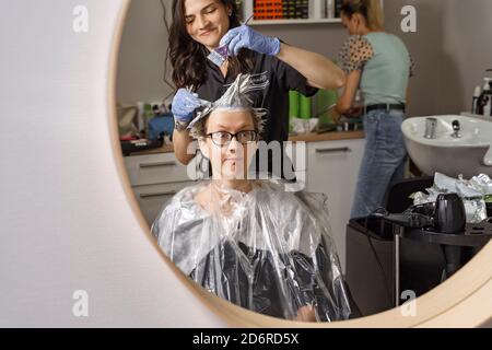 the hairdresser brightens the hair of the client. young woman looks at the process through the mirror and reacts emotionally. a grimace of fear and su Stock Photo