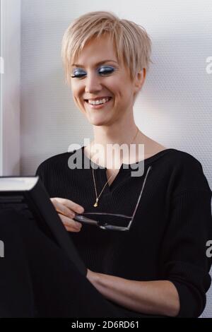 young blonde woman is reading a book by the window and smiling. glasses in hand Stock Photo