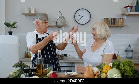 Elderly grandparents dancing while cooking together in kitchen. Senior woman and man making a funny dance with strainers. Healthy joyful mature family lifestyle concept Stock Photo