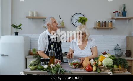 Senior grandparents in kitchen. Elderly woman and man cooking salad with fresh vegetables. Funny grandfather joking on grandmother. Putting a lettuce about her head. Healthy mature family lifestyle Stock Photo