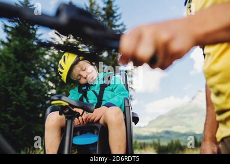 Unrecognizable father with sleeping small son cycling outdoors in summer nature. Stock Photo
