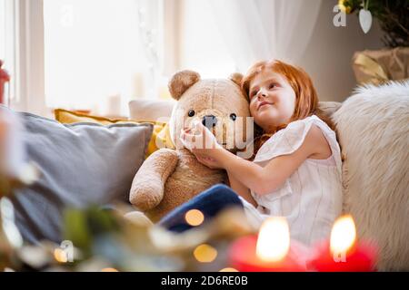 Small girl sitting on sofa indoors at home at Christmas, hugging teddy bear.