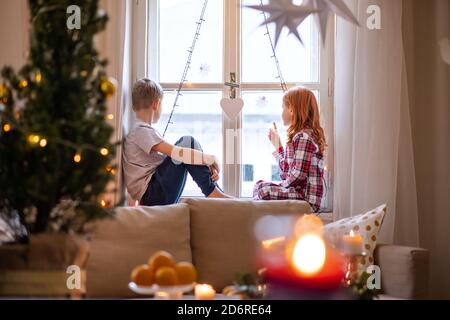 Small girl and boy in pajamas indoors at home at Christmas, sitting on window sill.