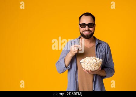 mage of a laughing emotional pleased young man in casual blue shirt eat popcorn wearing 3d glasses watch film isolated over yellow background. Stock Photo