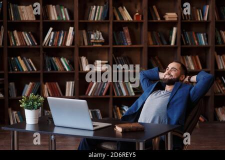 Young successful bearded businessman in a blue suit in the office library. businessman's Desk with laptop. time to relax Stock Photo