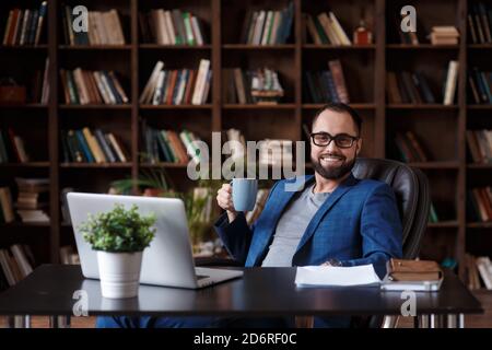 A young successful bearded businessman at his desk in the library office holds coffee in his hands and smiles. Stock Photo