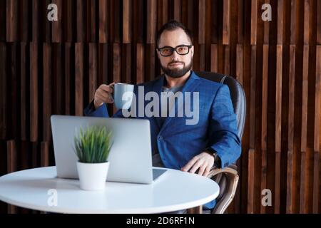 Young successful bearded businessman freelancer in office coworking. Happy man programmer developer in a white t shirt smiling and drinking coffee at Stock Photo
