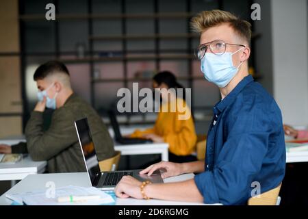 Young students with face masks at desks at college or university, coronavirus concept. Stock Photo