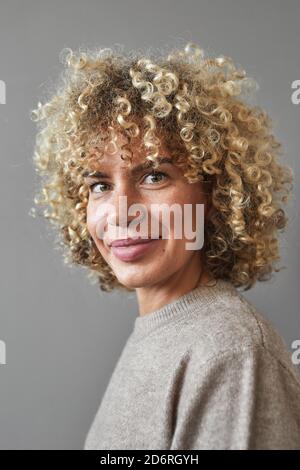 Vertical portrait of smiling curly haired woman looking at camera while posing against gray background, unique beauty concept Stock Photo