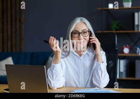 Smiling senior grey-haired beautiful businesswoman talking on the phone in office. Business portrait of attractive mature lady wearing eyeglasses Stock Photo