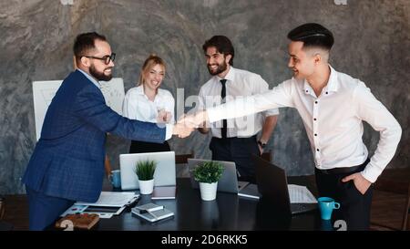 Glad to work with you Young modern men in smart casual wear shaking hands and smiling while working in the creative office Stock Photo