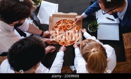 Happy diverse colleagues spend work break together eating takeaway delivery pizza in office, smiling multiracial employees have fun, laugh and chat Stock Photo