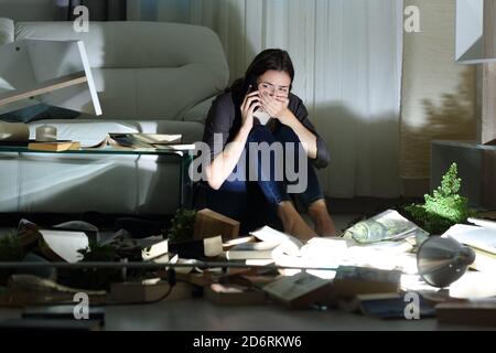 Scared home owner calling police after robbery sitting on the floor in the night with messy living room Stock Photo