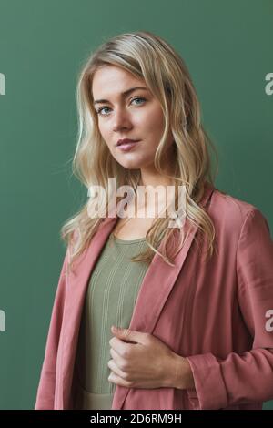 Vertical waist up portrait of elegant blonde woman wearing dusty pink jacket and looking at camera while posing against green background in studio Stock Photo