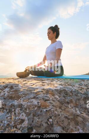 From below side view of concentrated serene young female sitting in Lotus pose with mudra gesture on top of rocky mountain and practicing yoga meditat Stock Photo