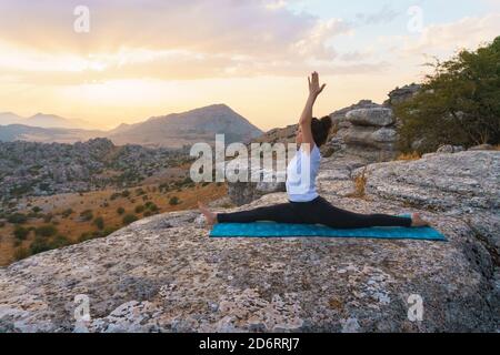 Back view of unrecognizable female performing front splits with arms raised while practicing advanced yoga on top of rock in highlands at sunset time Stock Photo