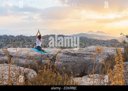 Back view of unrecognizable female performing front splits with arms raised while practicing advanced yoga on top of rock in highlands at sunset time Stock Photo