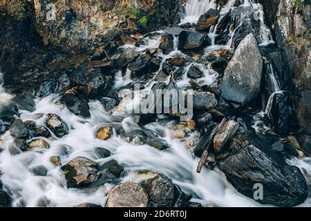 Long narrow waterfall of mountain river flowing over big stones between trees on cliffs in gorge Stock Photo