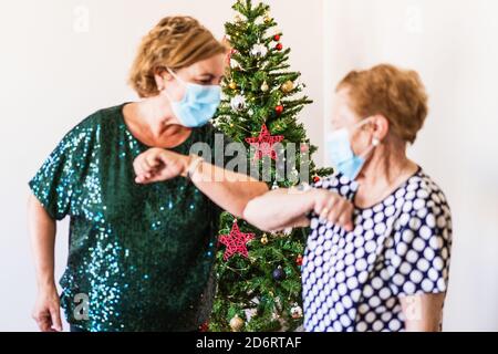 Mature women in smart casual outfits and face masks greeting by bumping elbows while standing near decorated shiny Christmas tree Stock Photo