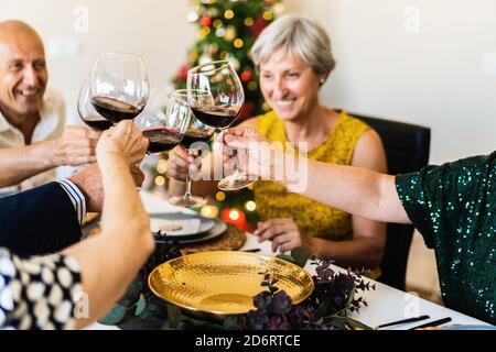 Happy mature friends gathering at festive table near shining Christmas tree and clinking wineglasses while celebrating Christmas eve together Stock Photo