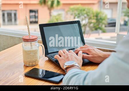 Crop faceless young male freelancer typing on laptop while working remotely in cafe sitting at table with smartphone and beverage Stock Photo