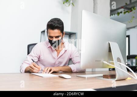 Anonymous concentrated businessman in mask taking note on paper near desktop computer while working on project Stock Photo
