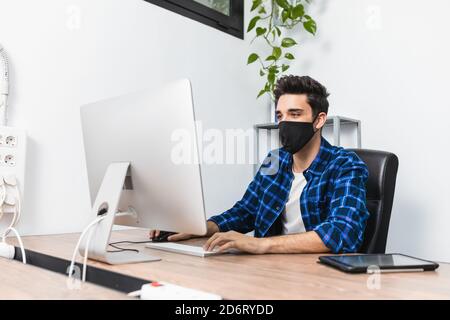 Unrecognizable young stylish male entrepreneur in face mask working on desktop computer in modern workspace Stock Photo