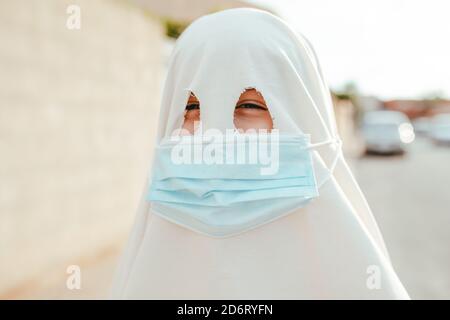 Anonymous spooky person wearing white ghost costume and medical mask standing on street and looking at camera during Halloween celebration Stock Photo