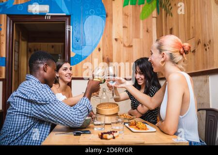 Group of cheerful multiethnic friends clinking wineglasses while having yummy dinner together in creative cafeteria Stock Photo