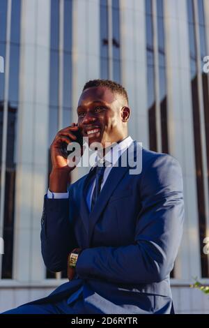 Low angle of happy successful well dressed black male manager in classy suit answering phone call while sitting against contemporary city building Stock Photo