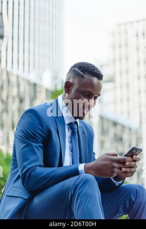 Low angle of happy successful well dressed black male manager in classy suit using smartphone while sitting against contemporary city building Stock Photo