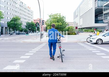 Back view of unrecognizable trendy male office worker in formal wear strolling on street with electric bicycle Stock Photo