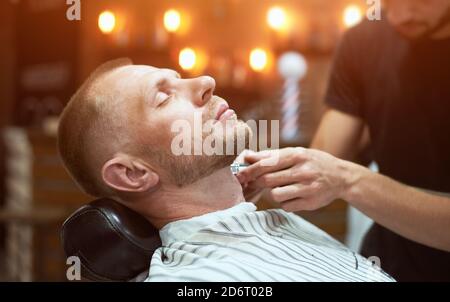 Unrecognizable blurred hairdresser is doing a perfect beard shape to his male client at barber shop, time to take care of oneself, relax, bright lights on the background Stock Photo