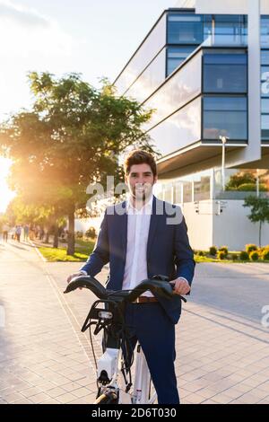 Trendy unshaven male office worker in formal wear strolling on street with electric bicycle while looking at camera Stock Photo