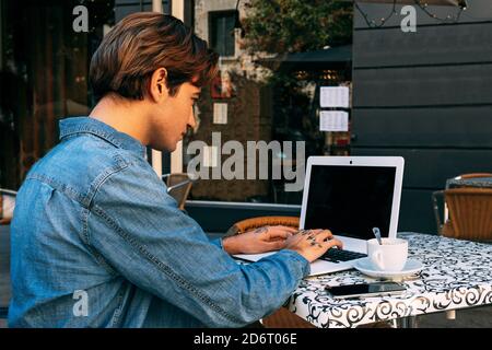 Side view of unrecognizable male freelancer with tattoo on hand sitting at table with cup of coffee and laptop while working on remote project in styl Stock Photo