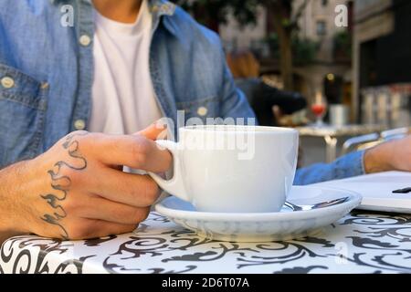 Crop anonymous male freelancer with tattoo on hand sitting at table with cup of coffee and laptop while working on remote project in stylish cafe