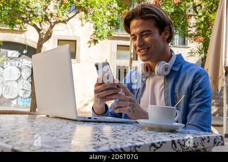 Positive young male remote worker in casual outfit with headphones on neck reading message on smartphone while sitting at table with laptop and cup of Stock Photo