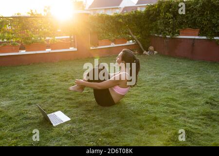 Side view of fit female athlete listening to online tutorial on laptop and doing abdominal crunches while balancing on grass during training at sunset Stock Photo